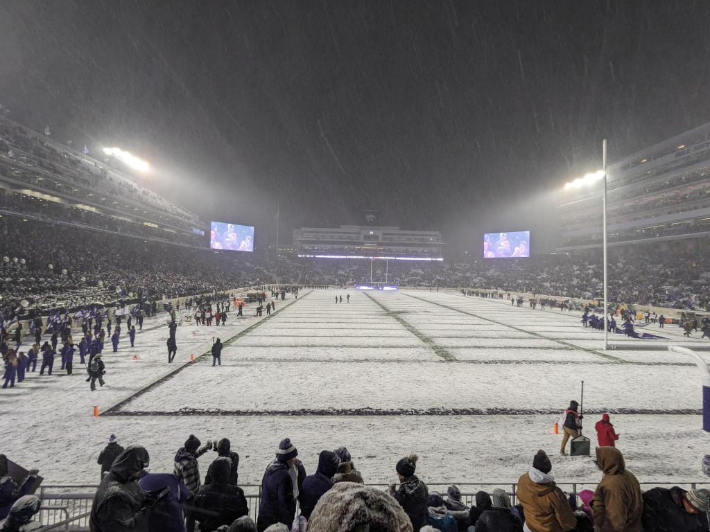 A snowy college football game between Kansas State and Iowa State, known as 'Farmageddon'. The scene depicts a night game with heavy snowfall, a packed stadium with fans bundled up in winter gear. The field is partly covered in snow, with players from both teams in action. Kansas State players are in purple and white uniforms, while Iowa State players are in red and gold. The snow adds a dramatic effect to the lights of the stadium, creating a chilly, winter atmosphere. The fans show mixed emotions, with some cheering and others showing disappointment.